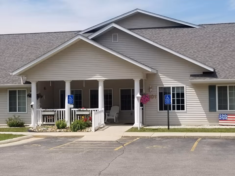 Front exterior view of a single-story building with beige siding and a gray shingled roof. The entrance features a covered porch supported by white columns, with a wheelchair accessible ramp and two handicap parking signs in front. There are windows on either side of the entrance and a small garden with flowers near the porch. An American flag decoration is visible on the right side of the building.
