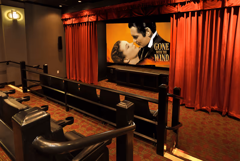 A cozy movie theater room with red curtains on the sides of a large screen displaying the movie poster for 'Gone with the Wind.' The room has two rows of black cushioned seats with armrests and cup holders, and the floor is carpeted in a red and orange pattern. Soft wall lighting adds to the warm ambiance.