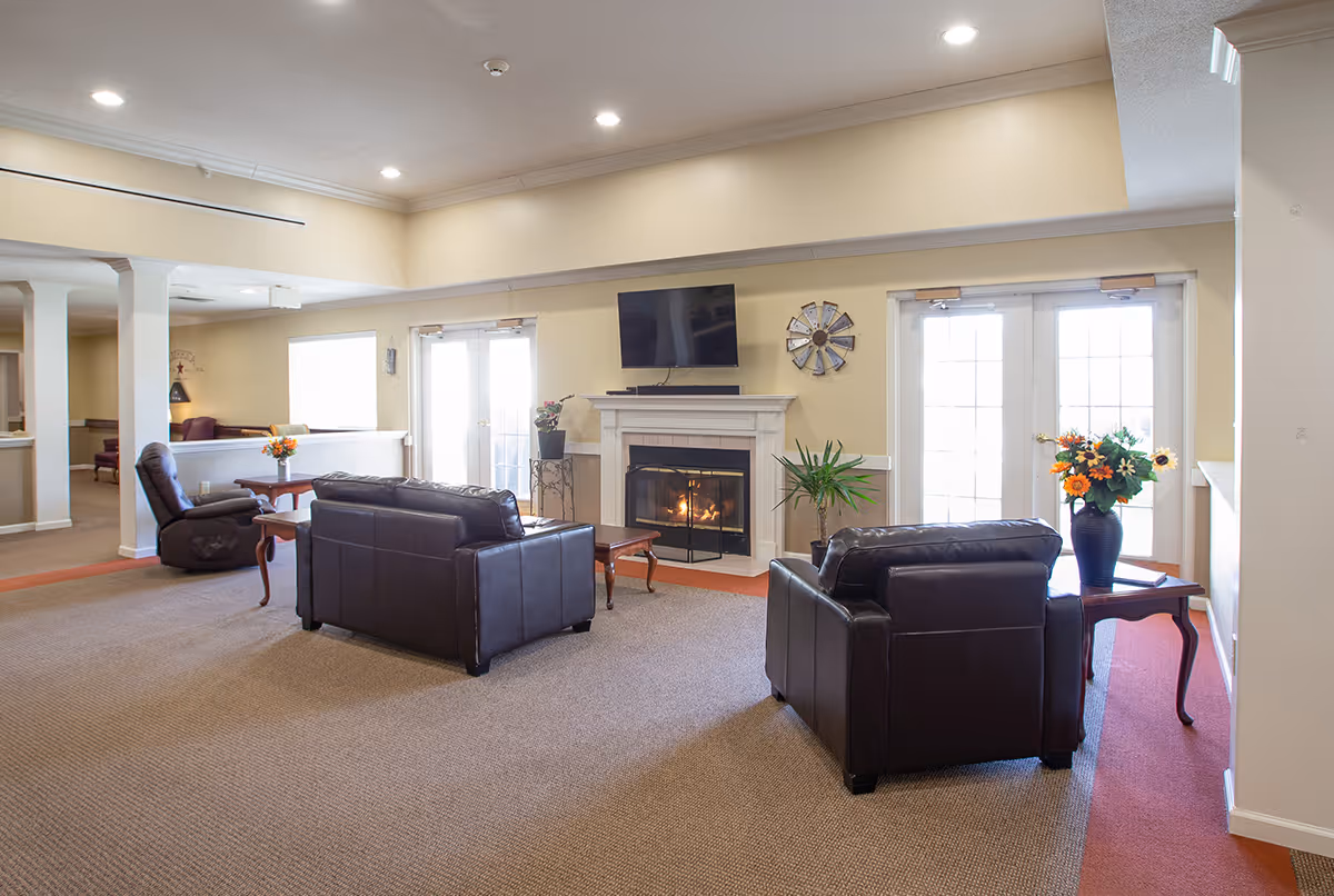 A bright and spacious senior living facility common area with beige walls and carpeted floors. The room features two dark brown leather sofas and a matching armchair arranged around a white fireplace with a fire burning inside. Above the fireplace is a mounted flat-screen TV and a decorative wall clock. There are two sets of French doors letting in natural light, and side tables with flower vases add a touch of color. The ceiling has recessed lighting, and there are plants placed near the fireplace.