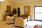 A cozy sitting area with two armchairs, a sofa, side tables, potted plants, framed artwork, and a glass door.