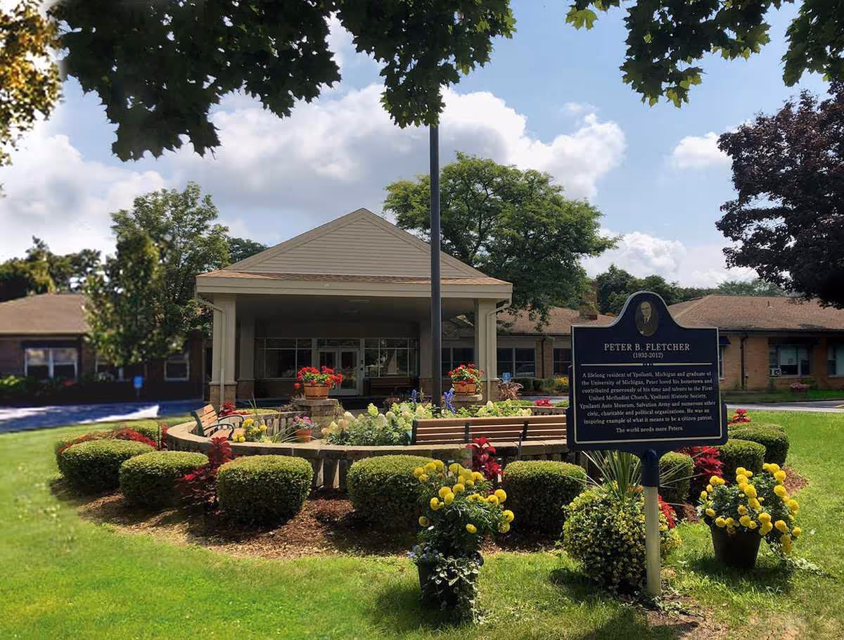 Exterior view of a senior living facility named Gilbert Residence with a well-maintained garden featuring trimmed bushes, colorful flowers, benches, and a commemorative sign for Peter B. Fletcher. The building has a covered entrance with columns and is surrounded by trees under a partly cloudy sky.