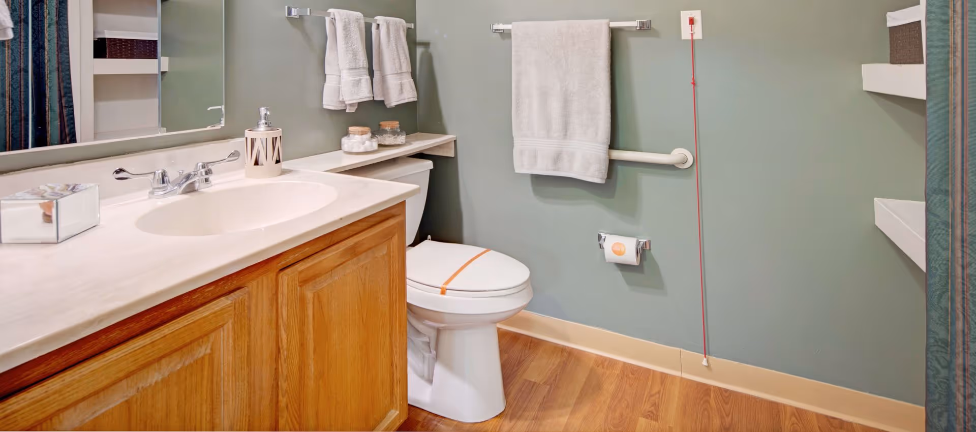 A clean bathroom with a wooden vanity and white countertop featuring a sink and faucet. Above the vanity is a large mirror. Next to the vanity is a white toilet with an orange strip across the seat. The walls are painted green and have two towel racks with white towels hanging. There is a grab bar on the wall next to the toilet and a red emergency pull cord. The floor is wood with a light baseboard.