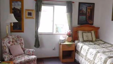 A small bedroom in a senior home with a bed, nightstand, lamp, window, and floral armchair.