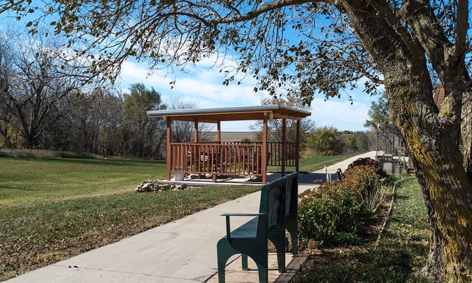 A peaceful outdoor area at Sabetha Manor featuring a paved walkway, a green bench, a wooden gazebo with a roof, surrounded by grass, trees, and shrubbery under a blue sky with some clouds.