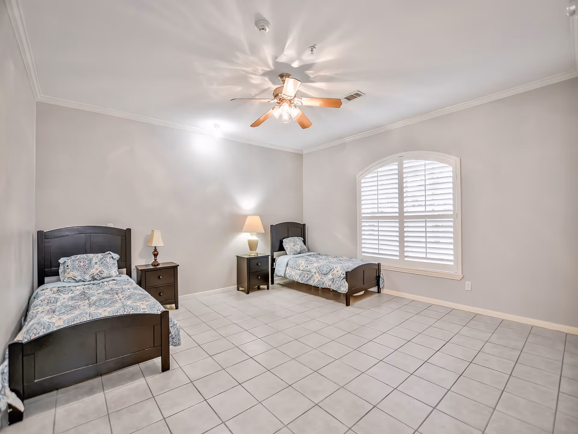 A spacious bedroom with two twin beds, nightstands and lamps, a ceiling fan, and a large arched window with shutters.