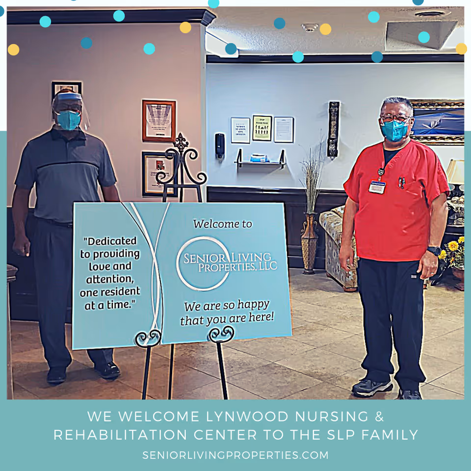 Two men wearing face masks standing indoors on either side of a sign that reads 'Welcome to Senior Living Properties, LLC. We are so happy that you are here!' The man on the left is wearing a face shield and dark clothing, while the man on the right is wearing red scrubs. The background shows a seating area with a couch, framed pictures, and a decorative vase.