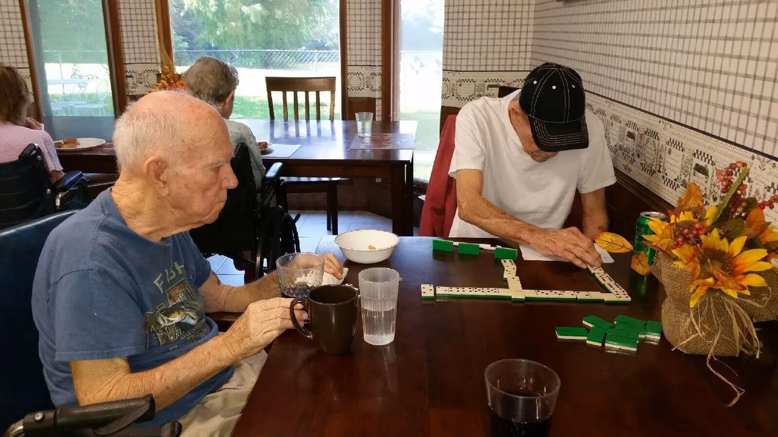 Two elderly men sitting at a wooden table playing dominoes in a room with patterned wallpaper and large windows showing greenery outside. One man is wearing a blue t-shirt and holding a coffee mug, while the other is wearing a white t-shirt and a black cap, focusing on the game. There is a vase with sunflowers and other autumn-themed decorations on the table.