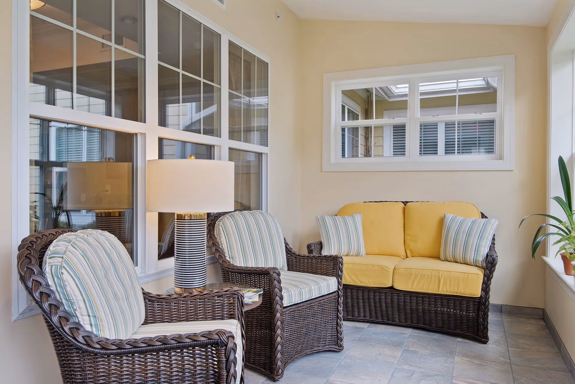 A cozy indoor seating area with wicker furniture including two armchairs with striped cushions and a loveseat with yellow cushions. A table lamp with a striped base is placed between the chairs. The room has large windows and a potted plant on the right side.
