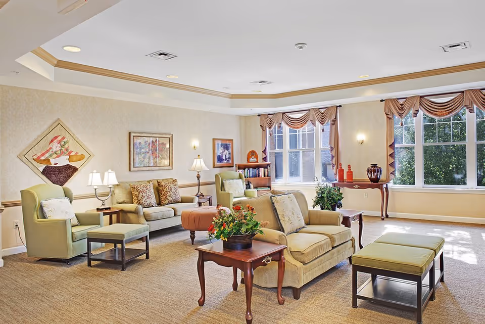 A bright and spacious living room with beige walls and carpeted floor. The room features multiple seating options including a green armchair with an ottoman, a beige loveseat, and a beige sofa with patterned cushions. There are several wooden side tables with lamps and a flower pot on one of the tables. Large windows with decorative curtains allow natural light to fill the room. The walls are adorned with framed artwork and a quilted wall hanging. A small bookshelf and decorative items are placed near the windows.
