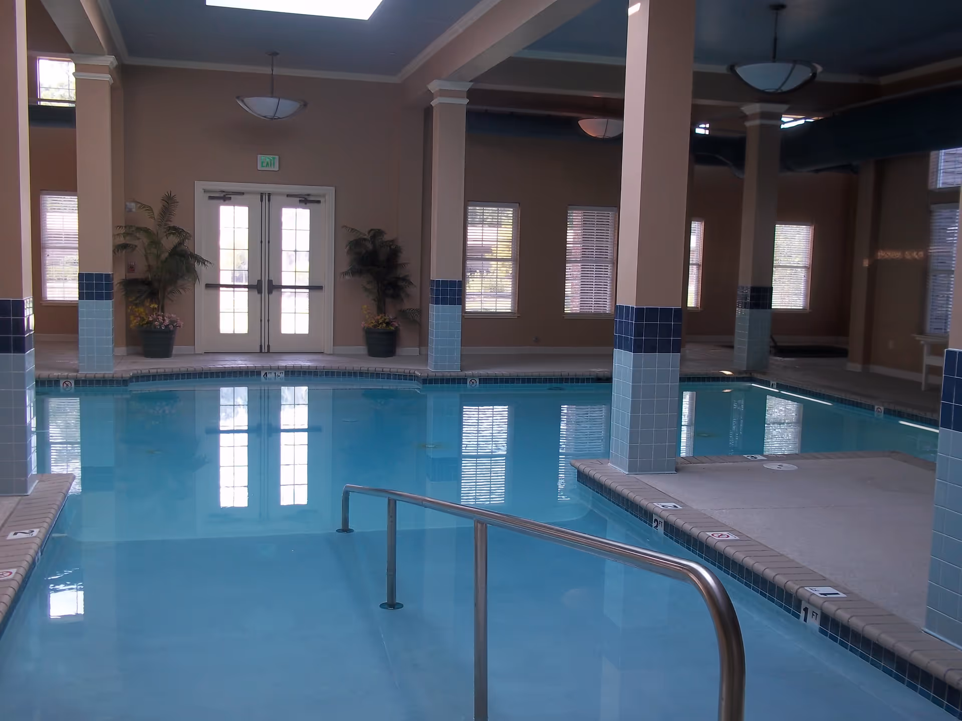 Indoor swimming pool with clear blue water, surrounded by beige walls and columns with blue tile accents. There are large windows with blinds and double glass doors at the far end. Two potted plants are placed near the doors. A metal handrail leads into the pool.