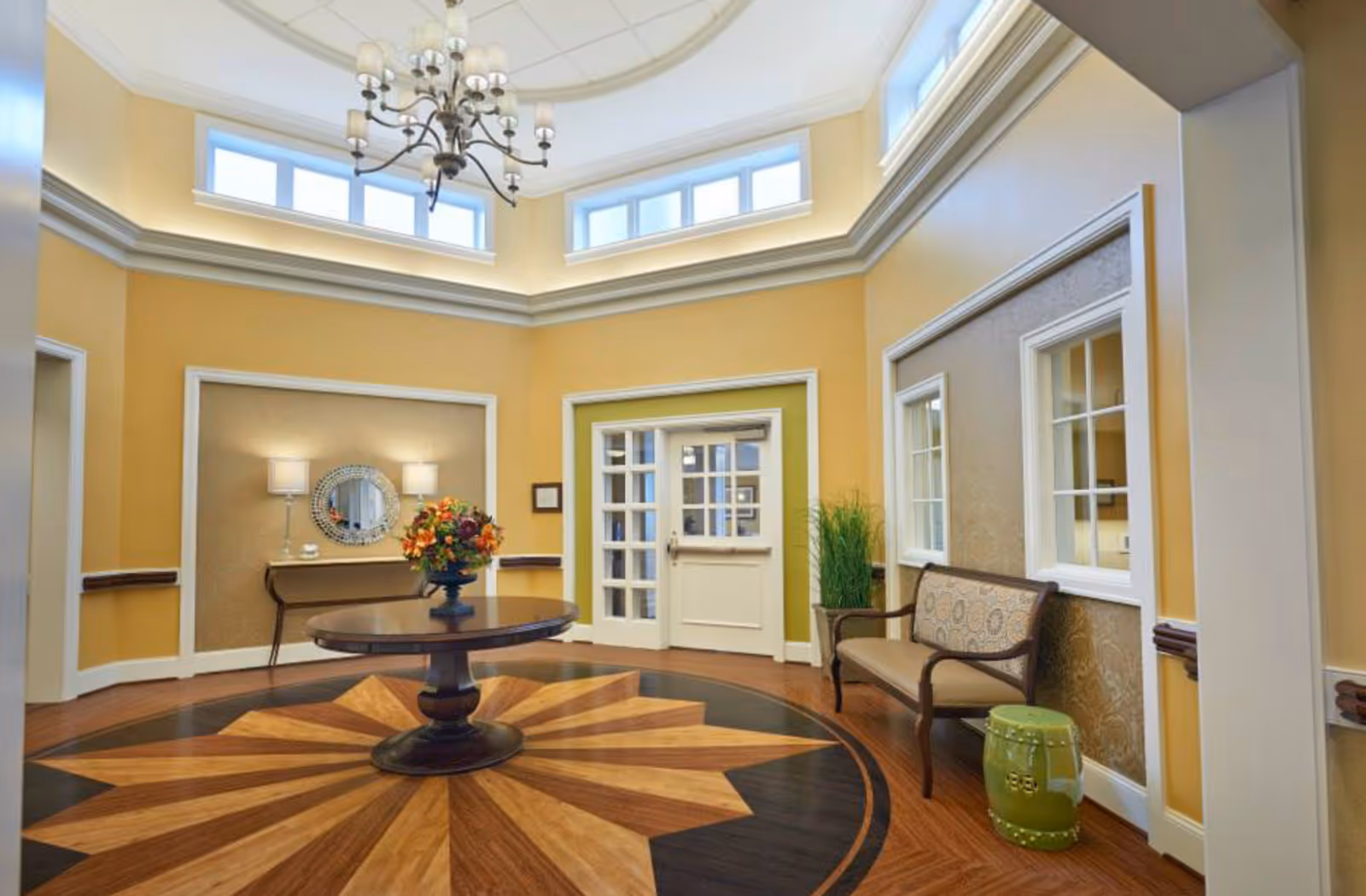 A bright and elegant interior room with yellow walls and high ceilings featuring a chandelier. The floor has a decorative wooden star pattern. In the center, there is a round wooden table with a floral arrangement. Against the walls, there is a console table with two lamps and a round mirror, a cushioned bench, and a green ceramic stool. Windows and a door with glass panes are visible on the walls.