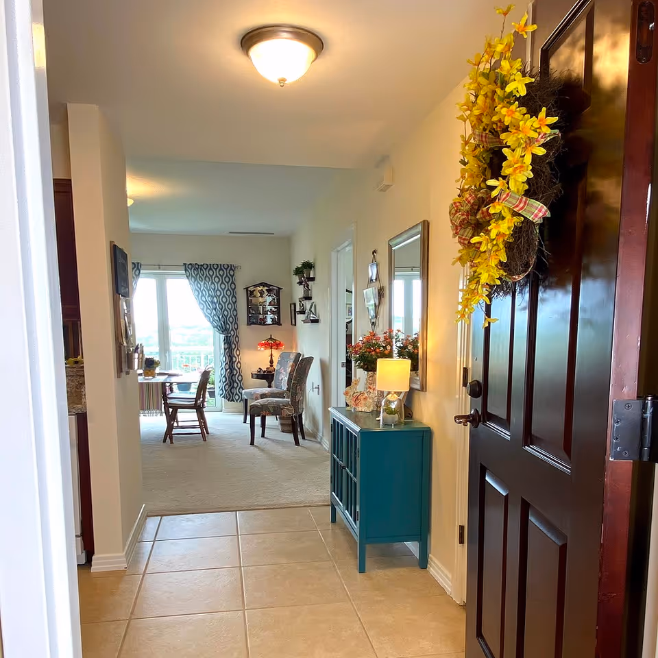 View from an open dark wooden front door into a bright interior hallway with beige tiled floor leading to a carpeted living area. A teal console table with a lamp and flowers is against the right wall, above which hangs a mirror. The door has a decorative yellow flower wreath. In the background, there are two patterned chairs, a small table with a red lamp, and a dining table with chairs near a window with patterned curtains.