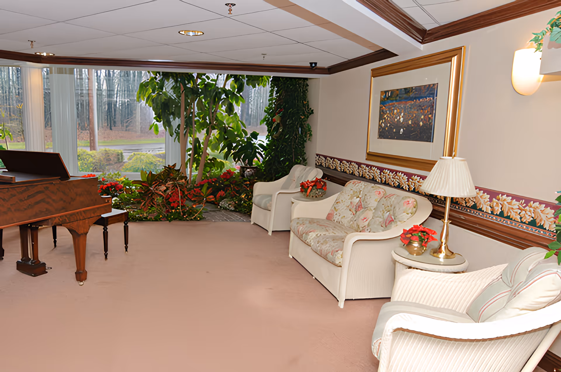 A cozy living room area with a floral-patterned loveseat, two armchairs, a side table with a lamp, and a wooden piano. Large windows reveal an outdoor view with trees and greenery. The room features beige walls with a floral wallpaper border and a framed painting.