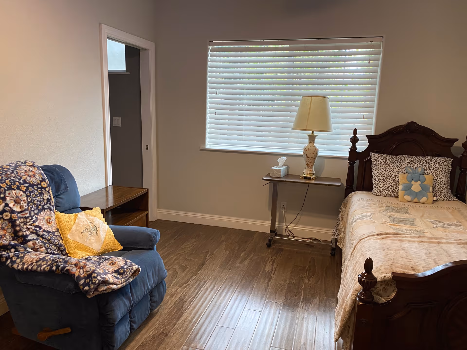 A cozy bedroom with a wooden bed featuring a patterned bedspread and decorative pillows, a small wooden side table with a floral lamp and tissue box, a blue recliner chair draped with a floral blanket and a yellow pillow, and a window with white blinds letting in natural light.