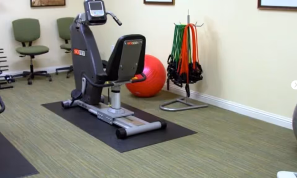 Indoor exercise room with a recumbent stationary bike on a black mat, two green office chairs with wheels against the wall, a red exercise ball, and a rack holding various resistance bands.