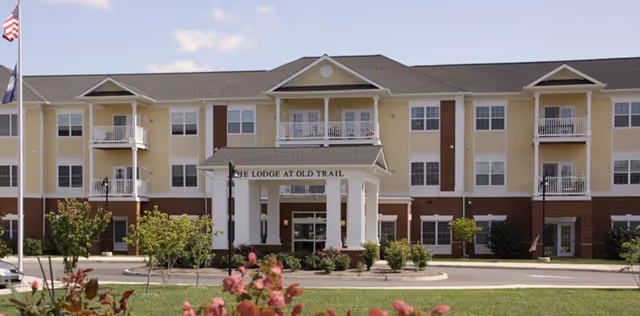 Front exterior view of The Lodge at Old Trail, a three-story building with balconies, a covered entrance with white columns, and landscaped greenery in front.