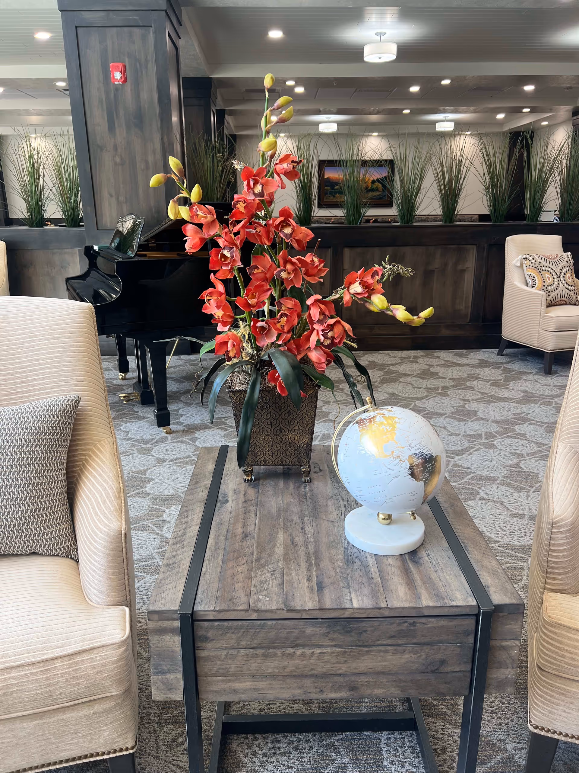A cozy senior living facility common area with two beige armchairs on either side of a wooden coffee table. On the table, there is a decorative globe and a vase with red flowers. In the background, there is a black grand piano, tall green plants, and a framed picture on the wall.