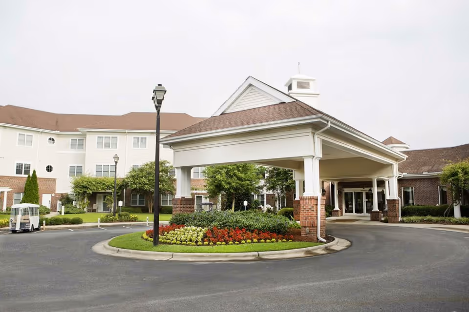 Front entrance of Cypress Glen Retirement Community featuring a covered driveway with brick pillars, landscaped flower beds, and a multi-story building in the background under an overcast sky.