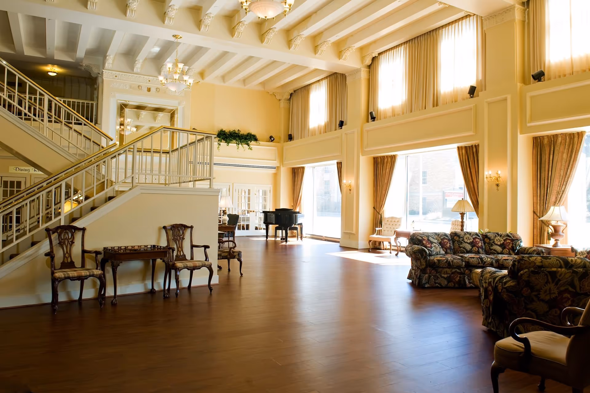 Spacious, sunlit senior living common room with high ceilings, a grand staircase, seating areas and a piano.