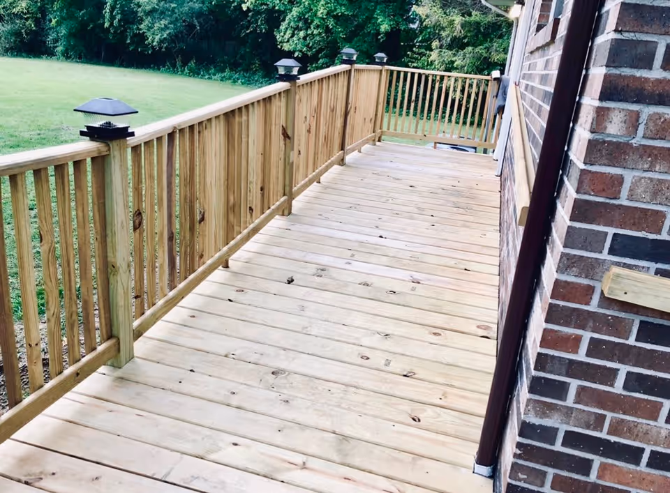 A wooden exterior deck with railing and small post lights running alongside a brick building overlooking a grassy yard.