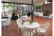 Enclosed sunroom/common seating area with white plastic tables and chairs, potted plants, a ceiling fan, and large windows.