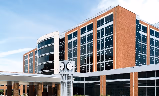 Exterior view of a modern multi-story hospital building with large windows and a clock tower in front under a blue sky.
