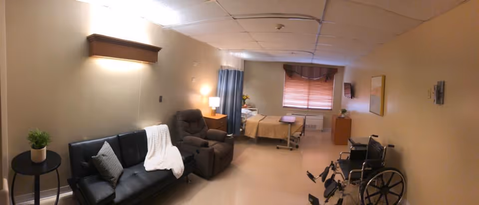A senior living facility room with a black sofa, a brown recliner chair, a hospital bed with beige bedding, a wooden nightstand with a lamp, a window with blinds and curtains, a small cabinet, and a wheelchair. The walls are beige and the floor is light-colored.