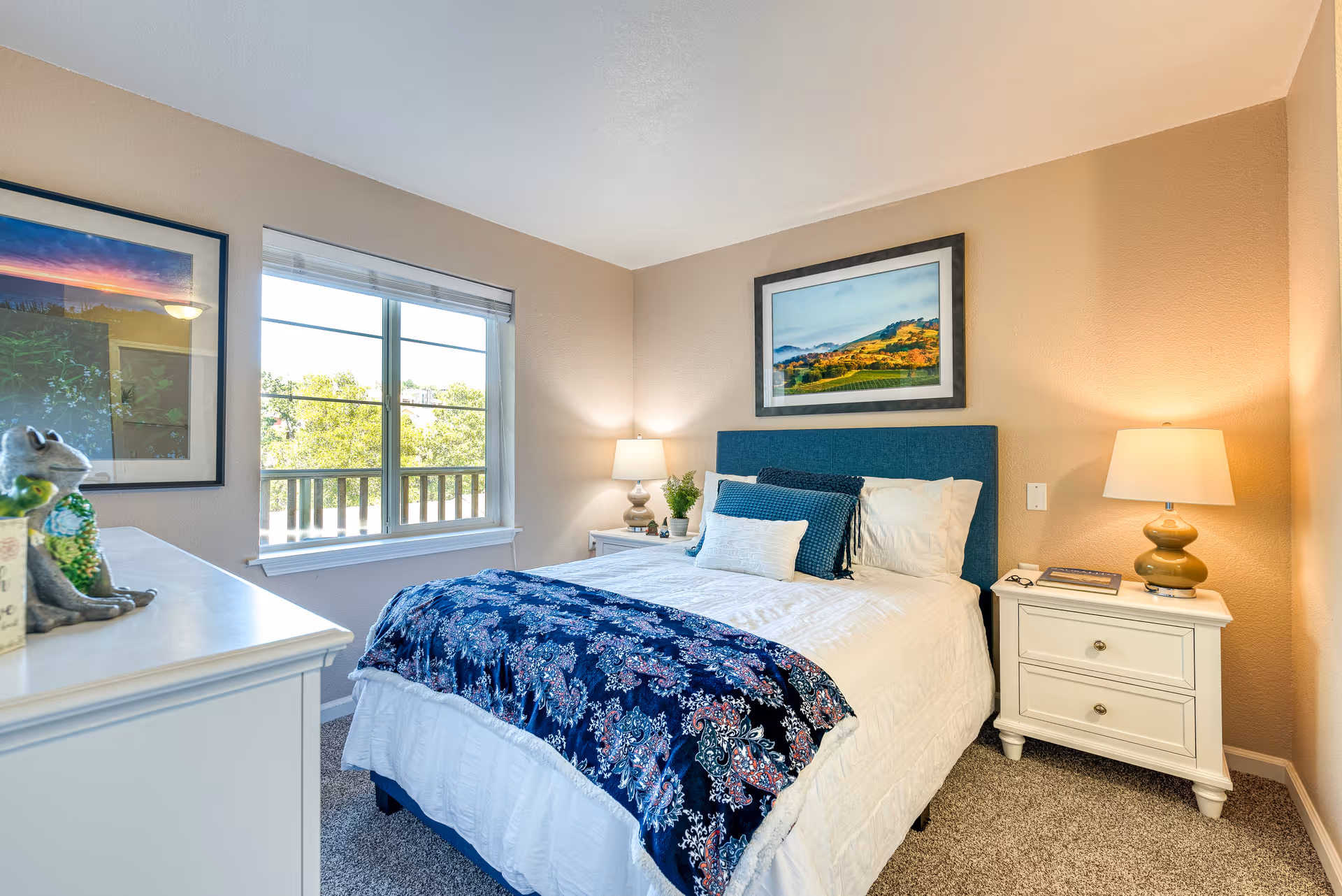 A cozy bedroom with a bed featuring a blue upholstered headboard, white bedding, and a blue patterned throw blanket. There are two white nightstands on either side of the bed, each with a lamp. A window with a view of trees is on the left wall, and framed landscape artwork hangs above the bed. The room has beige walls and carpeted flooring.