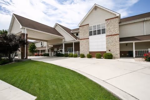 Exterior view of a senior living facility building with a covered entrance driveway, manicured green lawn, and clear blue sky with some clouds.