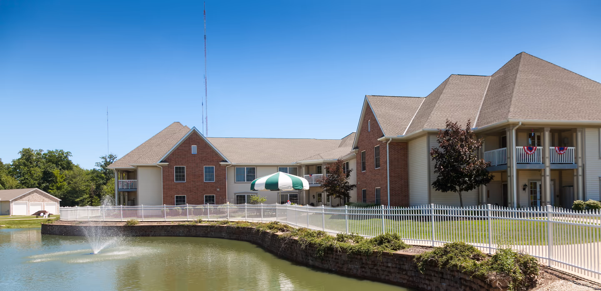 Exterior view of Mount Alverna Village senior living facility showing a large two-story building with brick and beige siding, a white fence, a pond with a water fountain, and a green and white striped umbrella near the building. The sky is clear and blue.