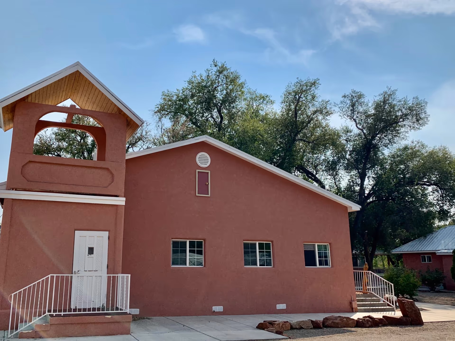 Exterior view of a single-story building with a reddish-brown stucco finish, three windows, a white door with a small porch and railing, and a small tower-like structure with a peaked roof. There are trees in the background under a clear blue sky.