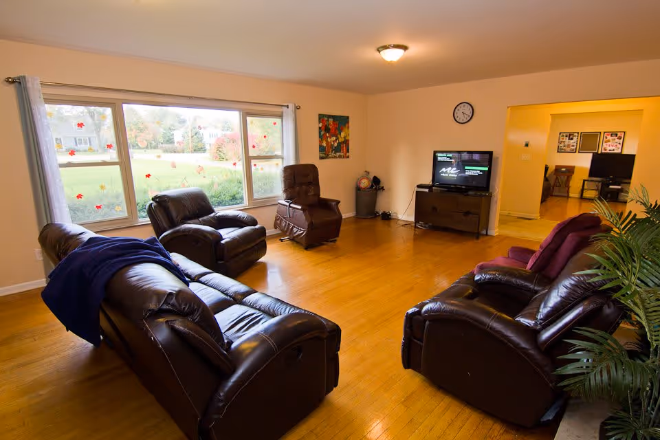 A spacious living room with wooden flooring, featuring three dark brown leather recliners and a matching sofa. A large window with light curtains lets in natural light and shows a green outdoor area. A small TV is placed on a wooden stand against the wall, with a clock above it. There is a colorful painting on the wall and a potted plant in the corner.