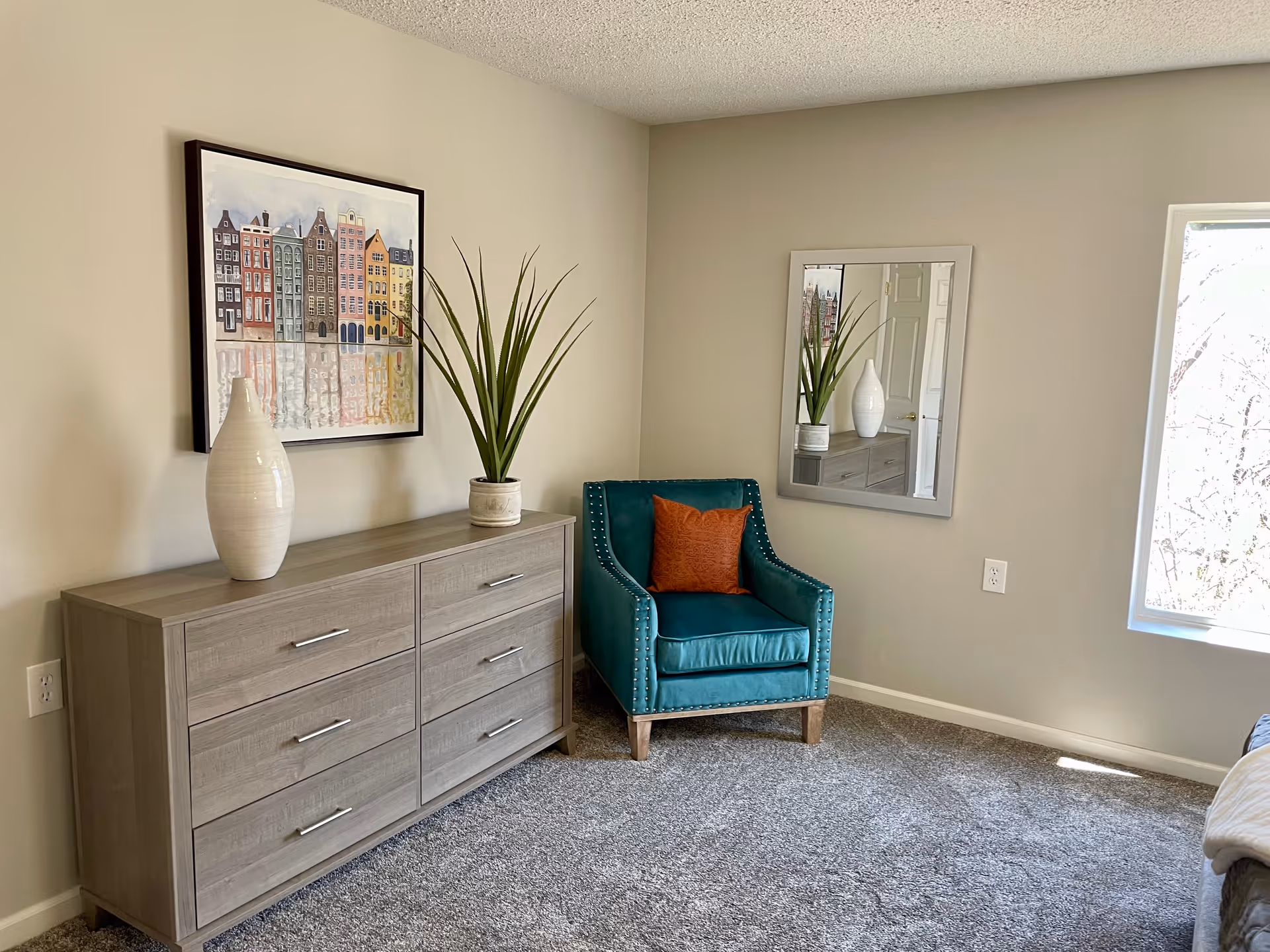 A corner of a bedroom featuring a gray wooden dresser with six drawers, decorated with a white vase and a potted plant. Above the dresser hangs a framed artwork of colorful buildings reflected in water. Next to the dresser is a teal upholstered armchair with a rust-colored pillow. A rectangular mirror is mounted on the adjacent wall, reflecting part of the dresser and plant. A window on the right side lets in natural light, illuminating the carpeted floor.