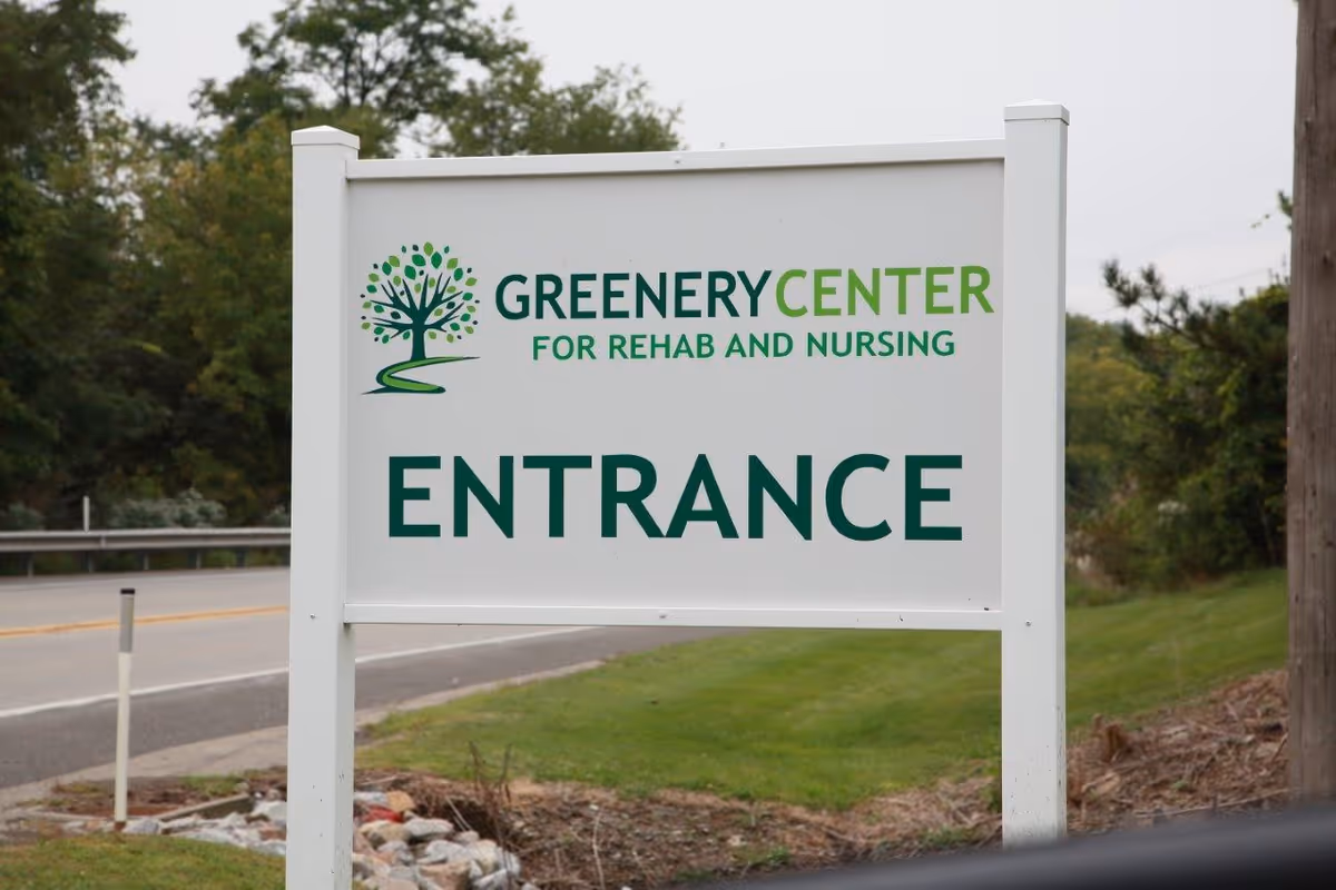 A white signboard with green text and a tree logo that reads 'Greenery Center for Rehab and Nursing ENTRANCE' located beside a road with trees and grass in the background.
