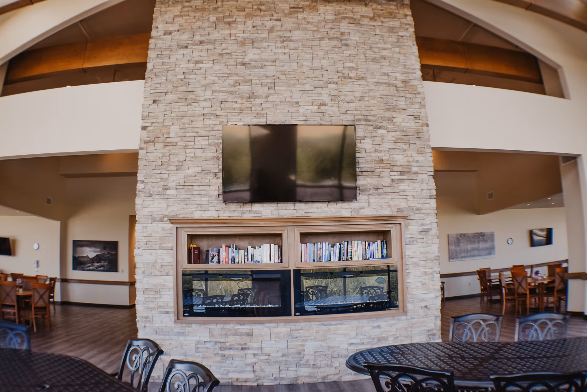 Communal lounge with a floor-to-ceiling stone fireplace wall, mounted TV, bookshelf and dining tables in the background.
