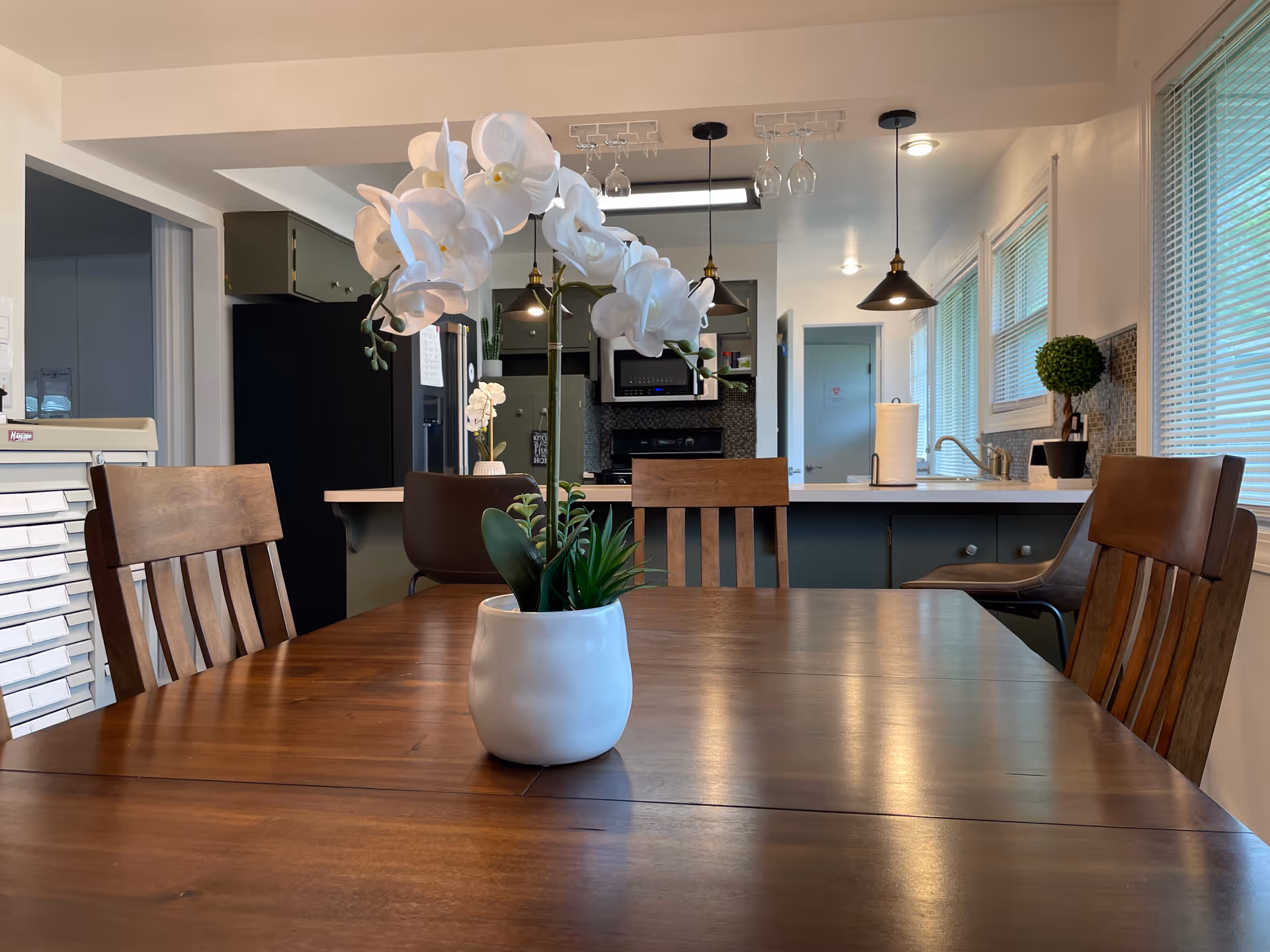 Dining table with a white orchid centerpiece and wooden chairs in front of a modern kitchen with pendant lights.