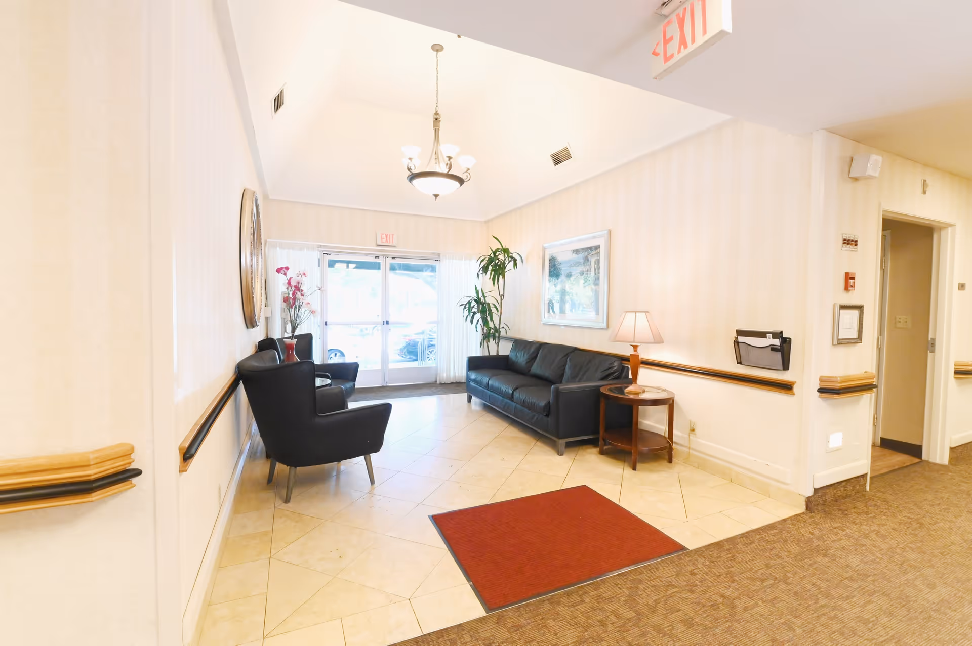 A bright and clean seating area in a healthcare facility with a black leather sofa, two black armchairs, a small round wooden side table with a lamp, a potted plant, and a framed picture on the wall. The area has tiled flooring with a red mat, beige walls with wooden handrails, and glass double doors leading outside. An exit sign is visible on the ceiling.