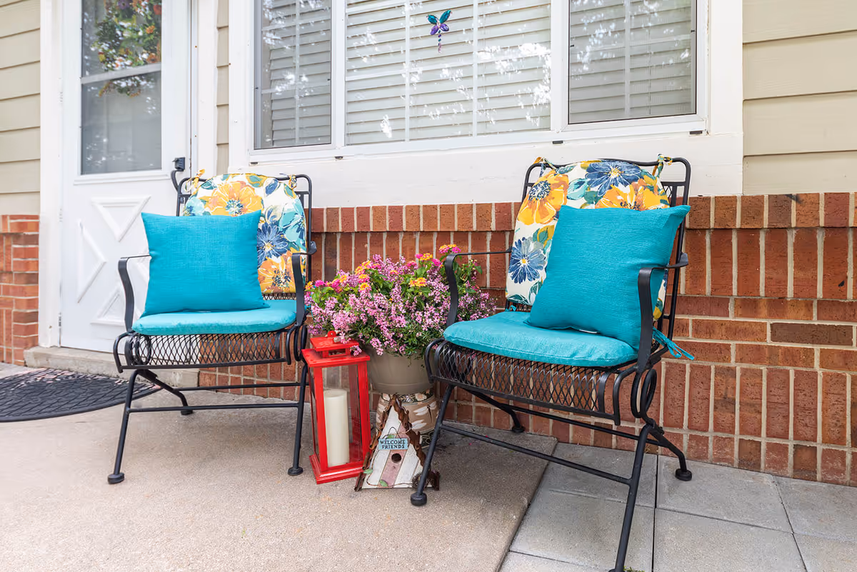 Two black metal chairs with turquoise seat cushions and floral back cushions are placed outside on a patio in front of a brick wall and window. Between the chairs is a pot of pink flowers, a red lantern with a white candle inside, and a small decorative birdhouse with a 'Welcome Friends' sign. A white door with a window is visible to the left.