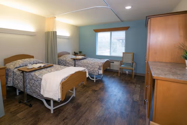 A healthcare facility room with two hospital-style beds, each with patterned bedding and a small overbed table. There is a window with blinds, a wooden chair, and a wooden cabinet with a plant on top. The floor is wood, and the walls are painted light blue and beige.