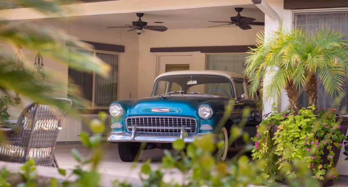 A vintage turquoise and white car parked under a covered patio area with ceiling fans, surrounded by greenery and potted plants.