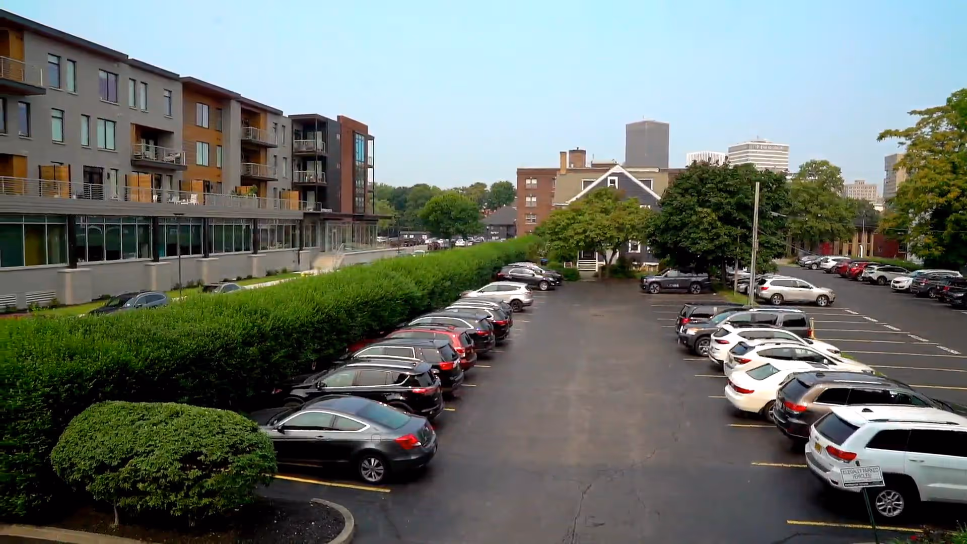Parking lot with multiple cars parked on both sides, adjacent to a multi-story residential building with balconies and large windows. Trees and bushes line the parking area, with a city skyline visible in the background under a clear sky.