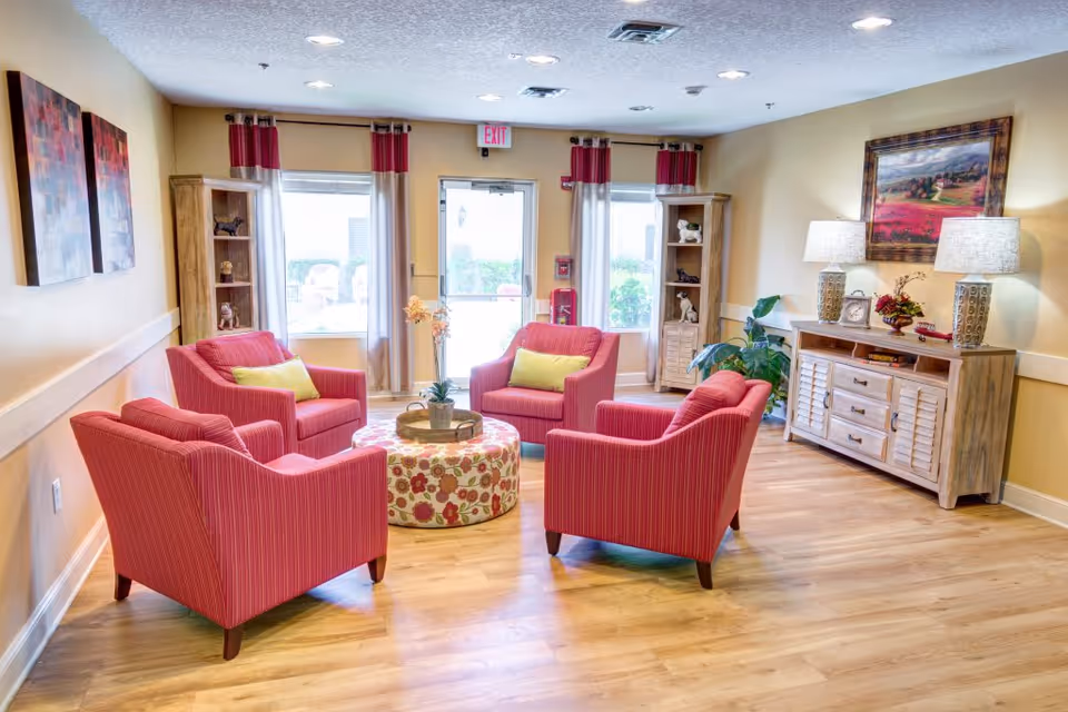 A bright communal lounge with four pink armchairs arranged around a floral ottoman, a wooden sideboard with lamps, and an exit door in the background.