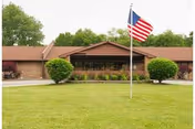 Single-story brick building with a brown roof, surrounded by green bushes and trees, with an American flag on a flagpole in the front lawn under a cloudy sky.