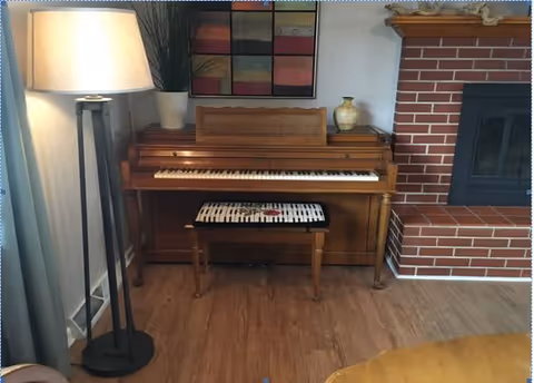 A wooden piano with a matching bench featuring a black and white patterned cushion is placed against a wall. To the left of the piano is a tall floor lamp with a white lampshade and a potted plant on top of the piano. Above the piano hangs a colorful abstract painting. To the right of the piano is a red brick fireplace with a black insert. The floor is wooden.