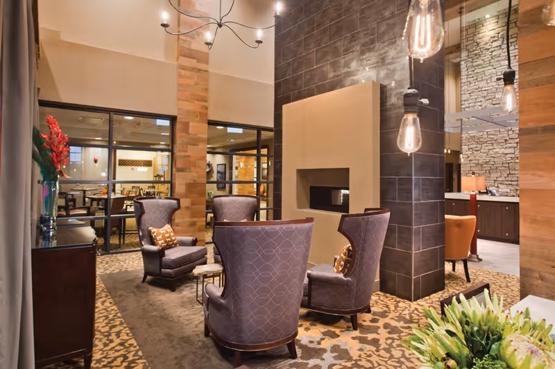 A cozy lounge area with four purple upholstered armchairs arranged around a modern fireplace with a dark tiled surround. The room features warm lighting with hanging Edison bulbs, patterned carpet, and wooden accents. In the background, there is a dining area visible through glass partitions.