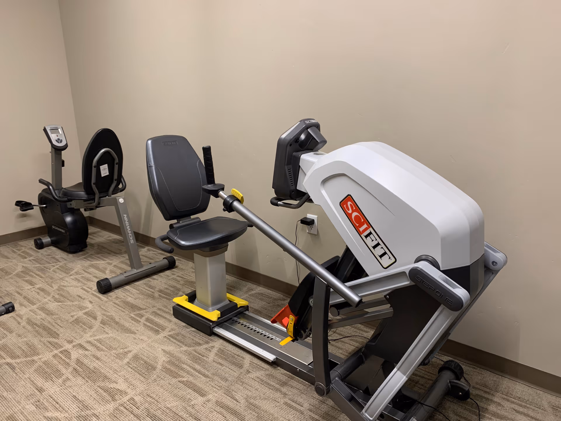 A small exercise room with two pieces of fitness equipment, including a SciFit seated stepper and a Schwinn recumbent bike, on a carpeted floor with beige walls.