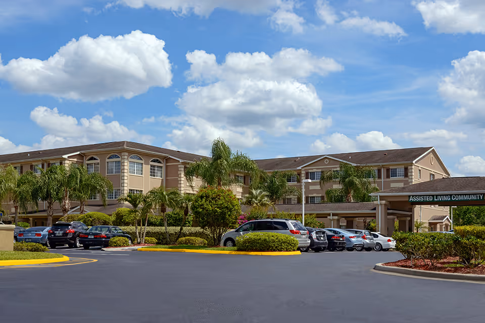 Front exterior of a three-story assisted living building with palm trees and parked cars under a blue sky.