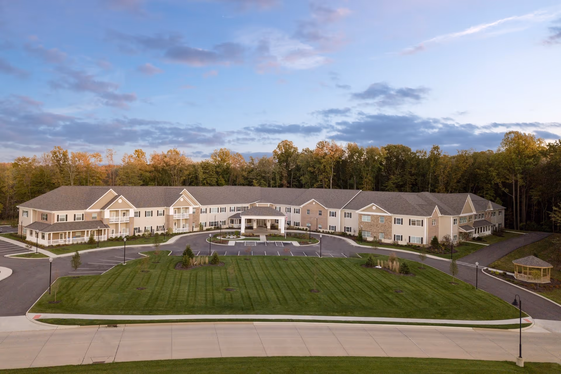 Aerial view of Danbury Brunswick senior living facility showing a large, two-story building with beige and stone exterior surrounded by a well-maintained lawn and trees in the background under a partly cloudy sky.