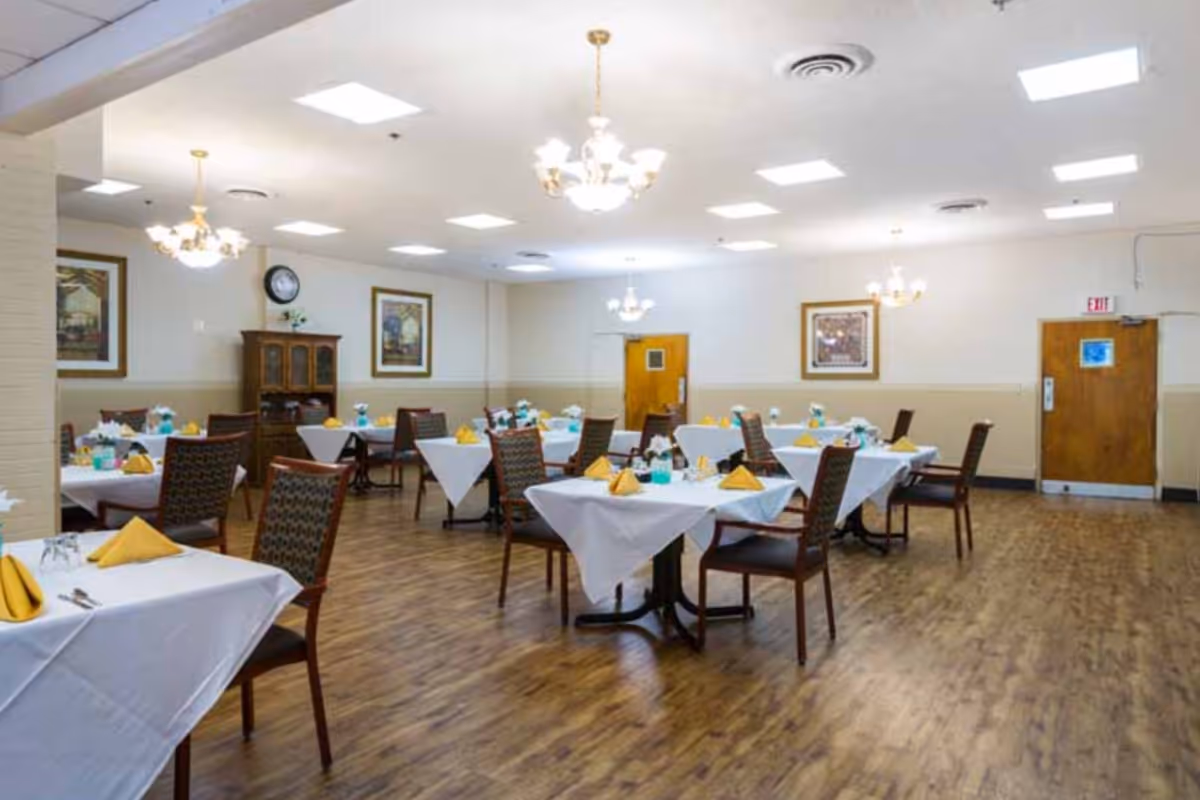 A dining room with multiple tables covered in white tablecloths, each set with yellow folded napkins, glassware, and floral centerpieces. The room has wooden floors, chandeliers hanging from the ceiling, framed artwork on the walls, and wooden doors.