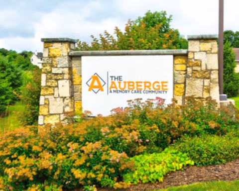 Stone sign for The Auberge, a memory care community, surrounded by green shrubs and plants with a partly cloudy sky in the background.